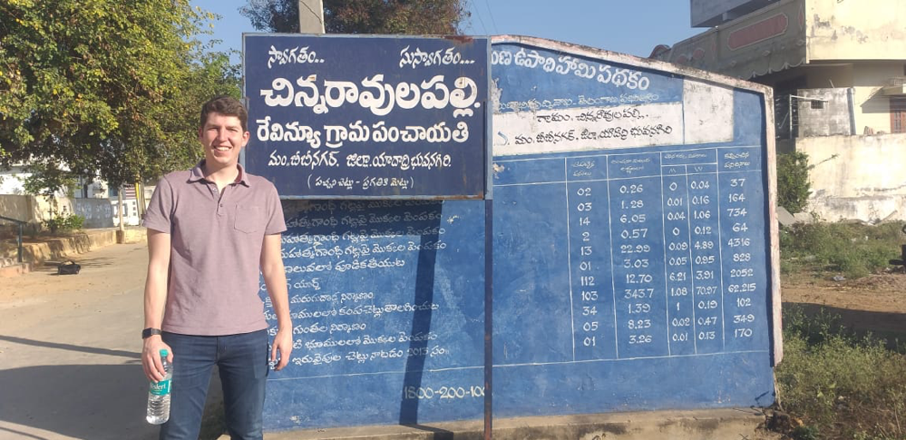 Amichai Feit stands in front of a board that conveys statistics and information about the National Rural Employment Guarantee Schemes (NREGS) program. Photo courtesy of Amichai Feit.